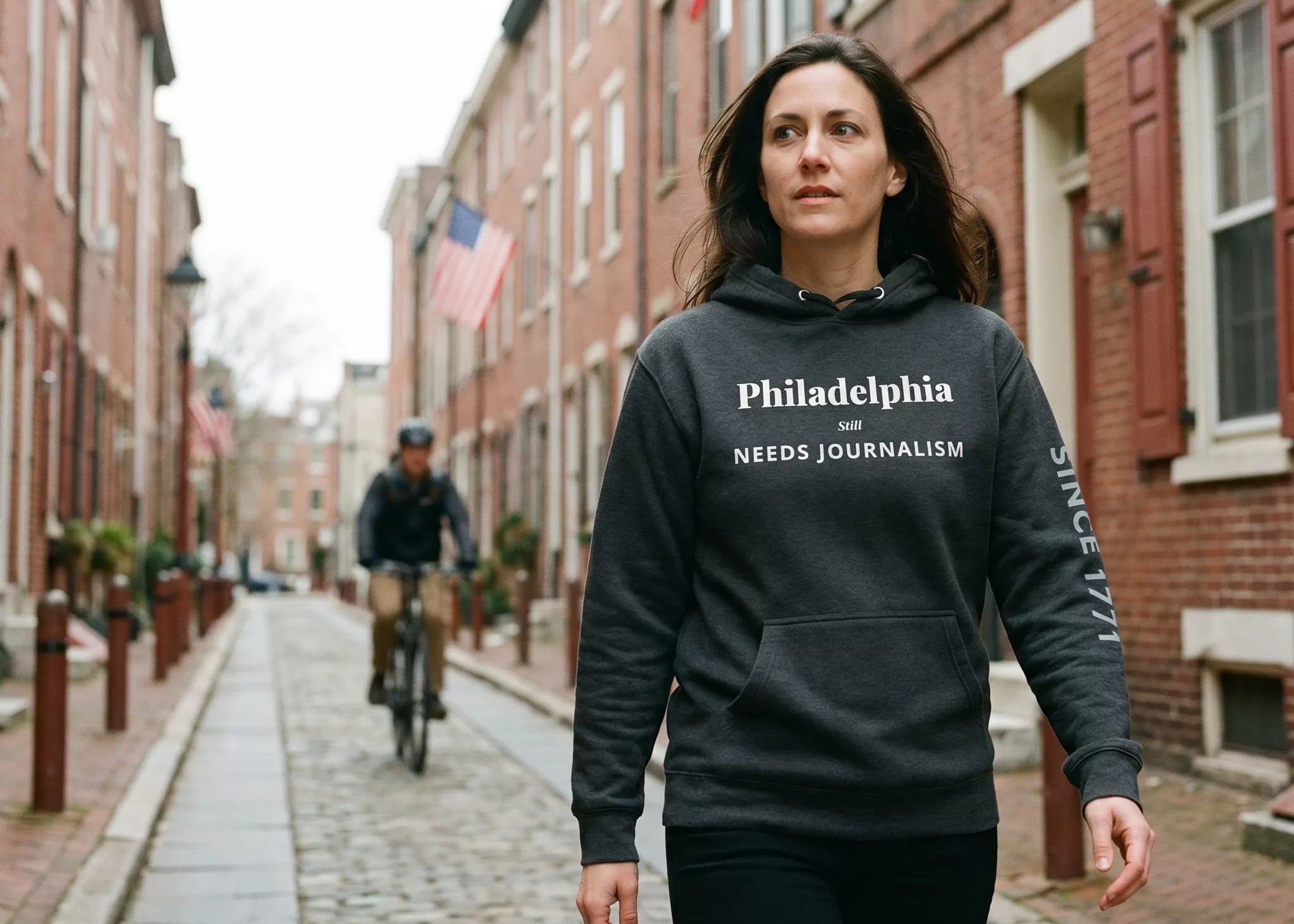 Woman wearing a hoodie with 'Philadelphia Needs Journalism' text walking down a street with American flags.